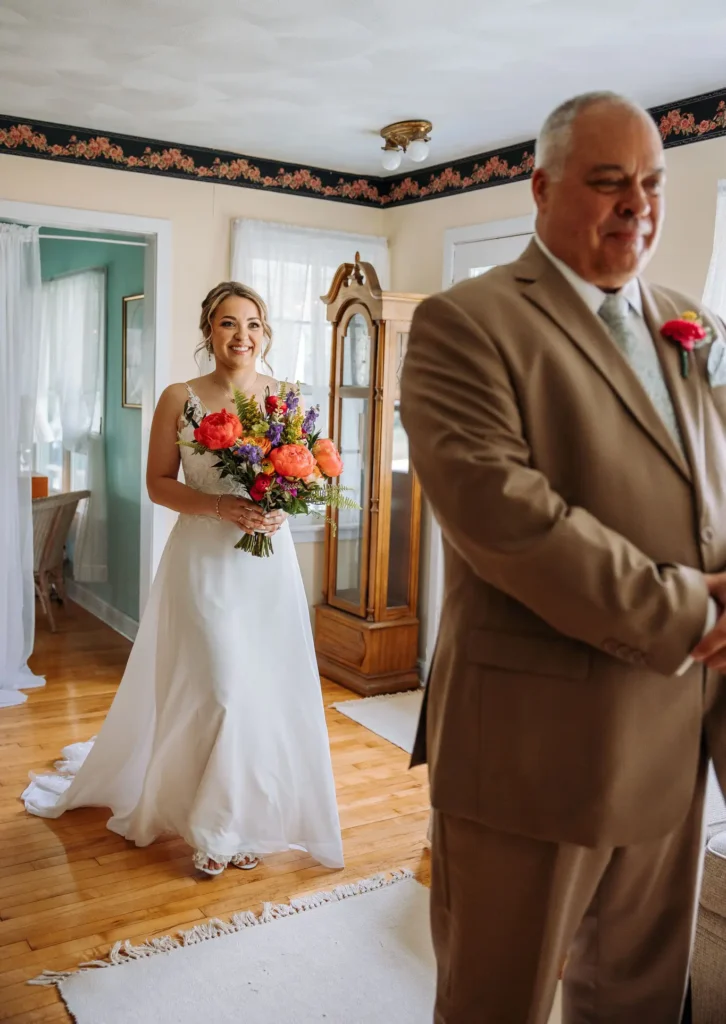 A bride in her dress stands in the bridal suite waiting for her father to turn around and see her at Old Coon Creek Wedding Venue in Beloit, Wisconsin