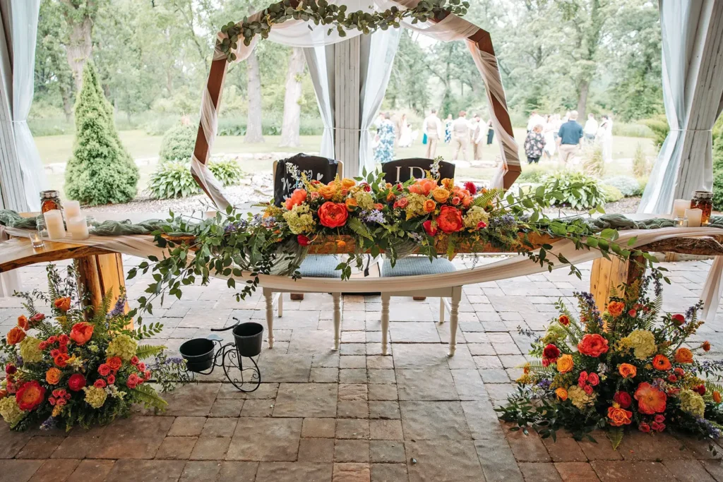 A bride and groom table is adorned with beautiful flowers while the wedding party stands in the distance at Old Coon Creek Wedding Venue in Beloit, Wisconsin